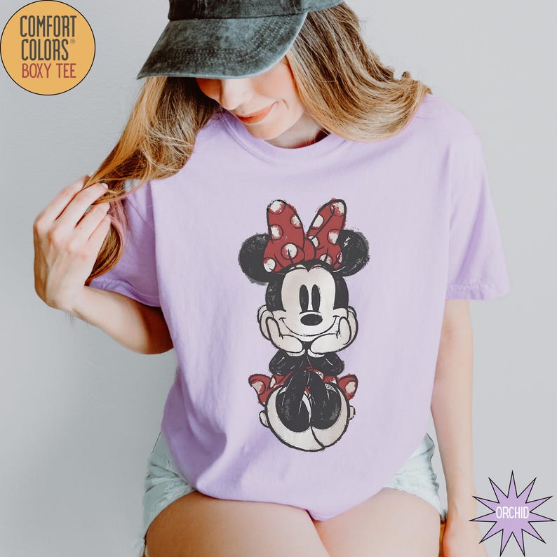 May include: A light purple Comfort Colors boxy tee featuring a distressed Minnie Mouse graphic. Minnie is depicted in black, red, and white, with a red polka dot bow. The shirt has short sleeves and a relaxed fit. The text "COMFORT COLORS" and "BOXY TEE" are printed on the upper left.
