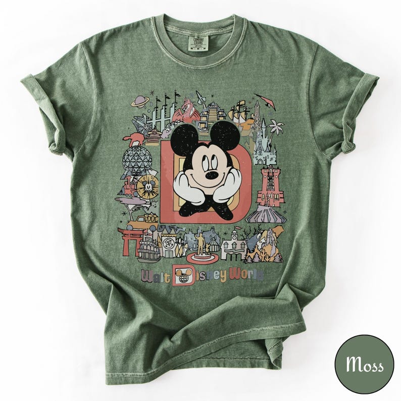 Puede incluir: Camiseta verde musgo con un gr&aacute;fico colorido de Mickey Mouse frente a la letra "D" con varios puntos de referencia de Disney World. Las palabras "Walt Disney World" est&aacute;n impresas en la parte inferior.