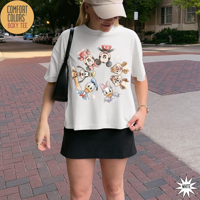 May include: White Comfort Colors Boxy Tee featuring a watercolor design of Disney characters including Mickey Mouse, Minnie Mouse, Donald Duck, Daisy Duck, Chip, and Dale. The shirt has short sleeves and a relaxed fit.