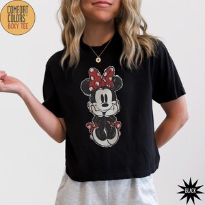 May include: Black boxy tee featuring a distressed graphic of Minnie Mouse with a red polka dot bow. The shirt has the words "Comfort Colors Boxy Tee" printed on the upper left side. The word "Black" is printed on the lower right side.