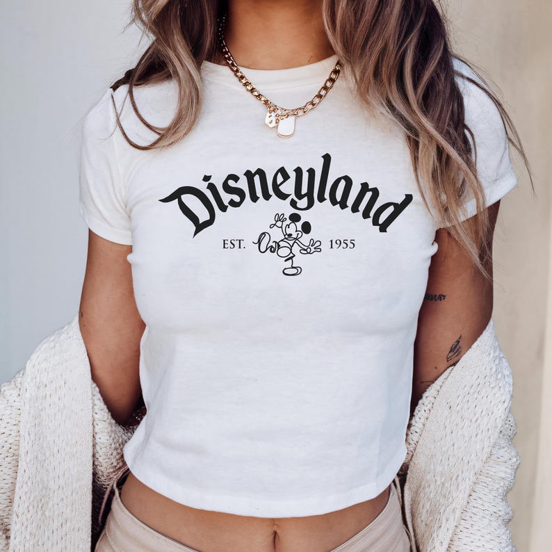 May include: A cream-coloured crop top featuring the word "Disneyland" in black script. Below the word is a Mickey Mouse graphic and the text "EST. 1955". The top has short sleeves and a round neckline.