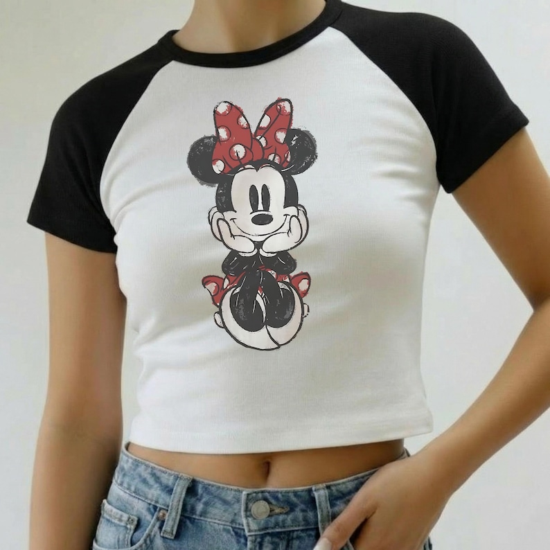 May include: White crop top with black raglan sleeves featuring a Minnie Mouse graphic. The graphic shows Minnie Mouse with a red polka dot bow and red polka dot skirt. The top is paired with blue jeans.