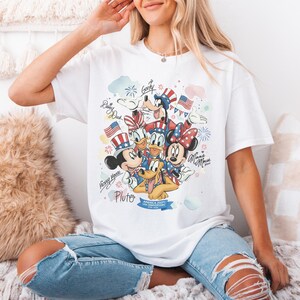 May include: White t-shirt featuring a colorful graphic of Disney characters Mickey Mouse, Minnie Mouse, Donald Duck, Daisy Duck, Goofy, and Pluto, celebrating the USA's 250th anniversary. The design includes fireworks, flags, and patriotic accents.