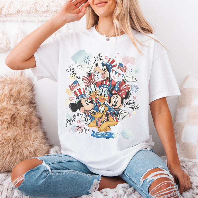 May include: White t-shirt featuring a colorful graphic of Disney characters Mickey Mouse, Minnie Mouse, Donald Duck, Daisy Duck, Goofy, and Pluto, celebrating the USA's 250th anniversary. The design includes fireworks, flags, and patriotic accents.