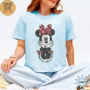 May include: Light blue Comfort Colors boxy tee featuring a distressed graphic of Minnie Mouse. Minnie is depicted in black, white, and red, with her signature polka dot bow. The shirt has short sleeves and a relaxed fit.