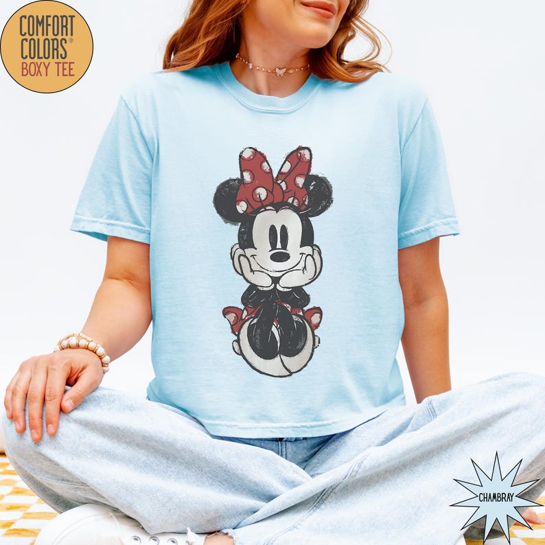 May include: Light blue Comfort Colors boxy tee featuring a distressed graphic of Minnie Mouse. Minnie is depicted in black, white, and red, with her signature polka dot bow. The shirt has short sleeves and a relaxed fit.