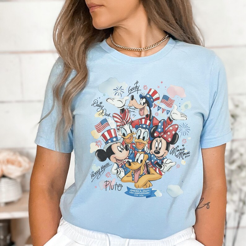May include: Light blue t-shirt featuring a colorful graphic of Disney characters Mickey Mouse, Minnie Mouse, Donald Duck, Daisy Duck, Goofy, and Pluto, with American flag-themed designs. The shirt includes text that reads "America 250th USA Anniversary 1776-2026".