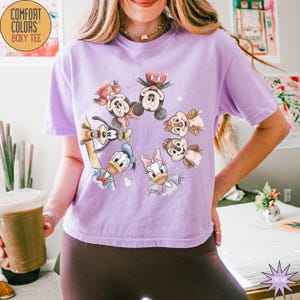 May include: A light purple Comfort Colors boxy tee featuring a circular graphic of Disney characters including Mickey Mouse, Minnie Mouse, Donald Duck, Daisy Duck, Goofy, and Chip and Dale. The shirt has short sleeves.