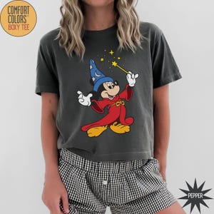 K&ouml;nnte beinhalten: Dunkelgraues Comfort Colors Boxy Tee mit einer Grafik von Mickey Mouse als Zauberlehrling. Mickey tr&auml;gt einen roten Umhang und einen blauen Hut und h&auml;lt einen Zauberstab mit Sternen. Das Shirt hat den Text "COMFORT COLORS BOXY TEE" in einem Kreis.