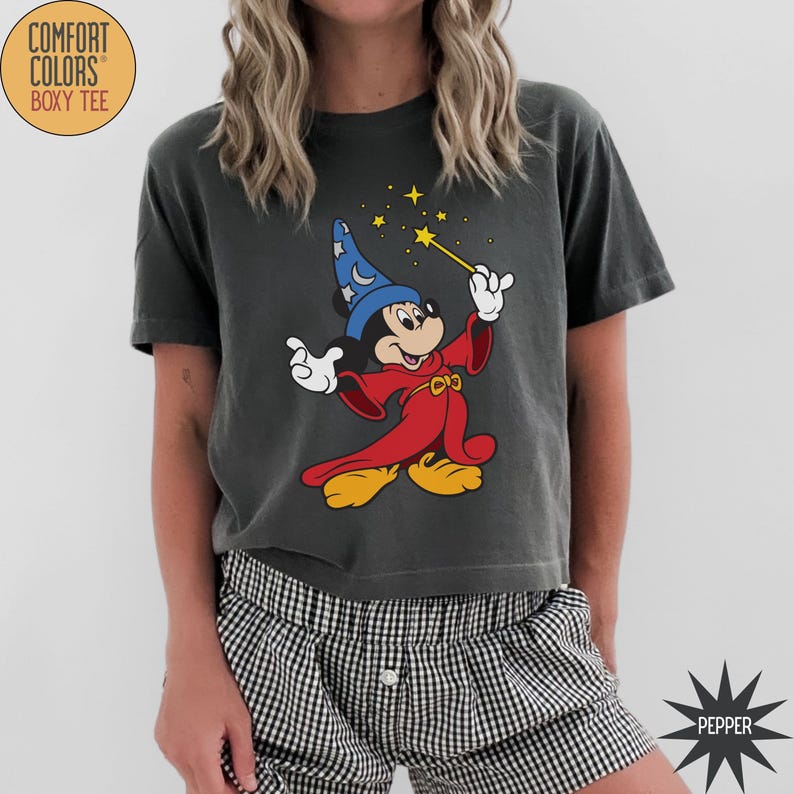 K&ouml;nnte beinhalten: Dunkelgraues Comfort Colors Boxy Tee mit einer Grafik von Mickey Mouse als Zauberlehrling. Mickey tr&auml;gt einen roten Umhang und einen blauen Hut und h&auml;lt einen Zauberstab mit Sternen. Das Shirt hat den Text "COMFORT COLORS BOXY TEE" in einem Kreis.