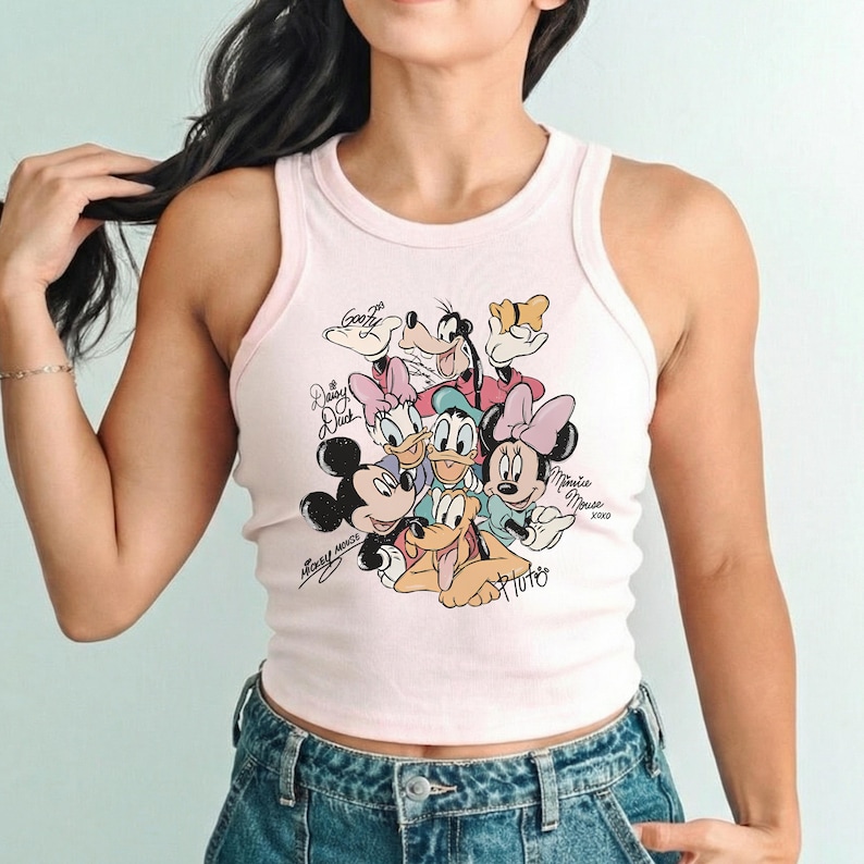K&ouml;nnte beinhalten: Ein hellrosa Tanktop mit einem farbenfrohen Aufdruck von Disney-Figuren, darunter Micky Maus, Minnie Maus und Donald Duck. Das Tanktop hat einen runden Ausschnitt und ein &auml;rmelloses Design. Der Text "Mickey Mouse" und "Minnie Mouse" ist ebenfalls sichtbar.