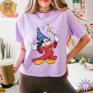 K&ouml;nnte beinhalten: Ein lila Comfort Colors Boxy Tee mit einem Cartoon-Mickey-Mouse-Zauberer-Design, der einen Zauberstab mit Sternen h&auml;lt. Das Design zeigt einen blauen Zaubererhut und rote Roben. Das T-Shirt hat eine lockere Passform.