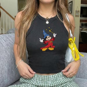 May include: Black tank top featuring a colorful graphic of Mickey Mouse dressed as a wizard, holding a magic wand. The sleeveless top has a round neckline and is made of a soft, stretchy material. The tank top is paired with green and white checkered pants.