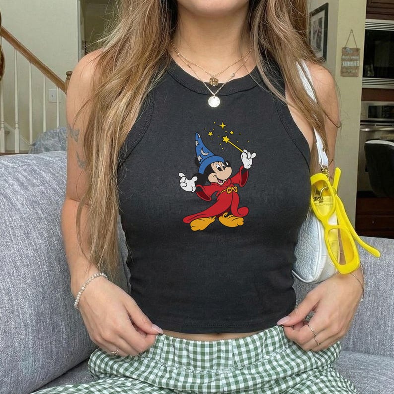 May include: Black tank top featuring a colorful graphic of Mickey Mouse dressed as a wizard, holding a magic wand. The sleeveless top has a round neckline and is made of a soft, stretchy material. The tank top is paired with green and white checkered pants.