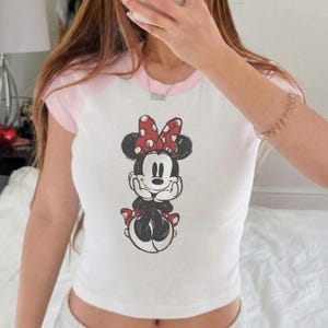 May include: White crop top with pink sleeves and collar featuring a Minnie Mouse graphic. The Minnie Mouse illustration has a red bow with white polka dots. The shirt is a casual, short-sleeved style.