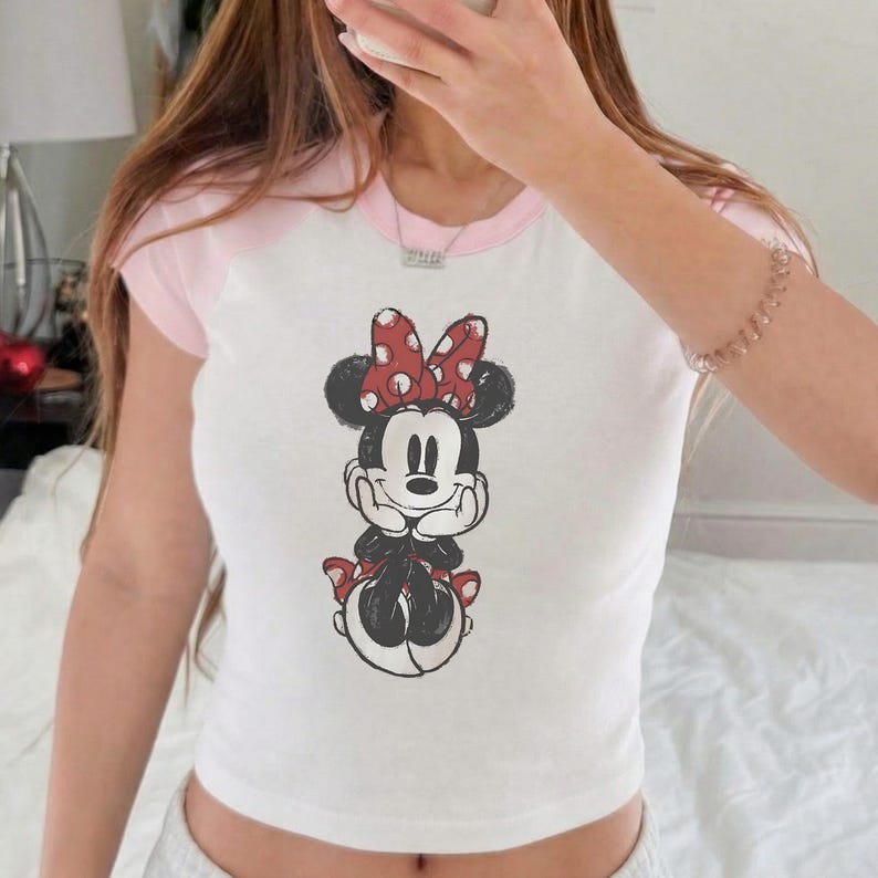May include: White crop top with pink sleeves and collar featuring a Minnie Mouse graphic. The Minnie Mouse illustration has a red bow with white polka dots. The shirt is a casual, short-sleeved style.