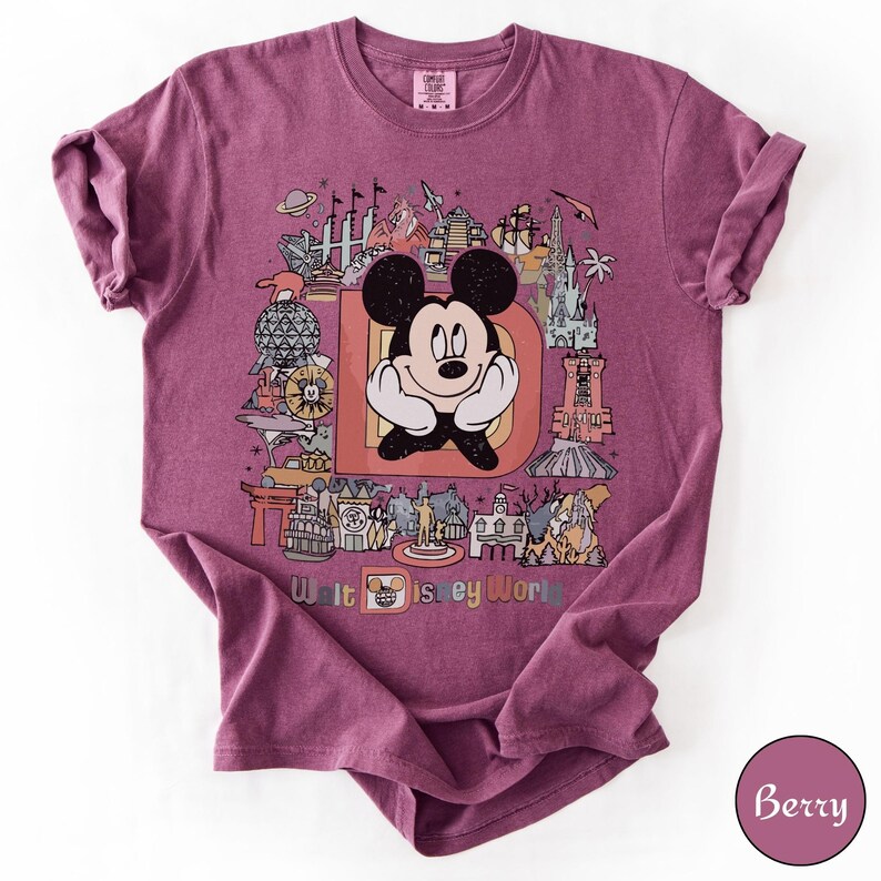 Puede incluir: Camiseta color bayas con un gr&aacute;fico colorido de Mickey Mouse dentro de una gran letra "D" con varios monumentos de Disney World. Las palabras "Walt Disney World" est&aacute;n impresas en la parte inferior. La camiseta tiene cuello redondo y mangas cortas.