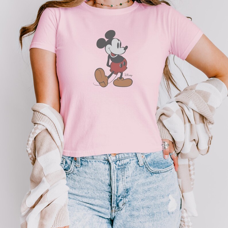 May include: A light pink t-shirt featuring a classic Mickey Mouse graphic. The cartoon character is depicted in black, red, and brown, walking with a smile. The shirt is short-sleeved and made of a soft material.