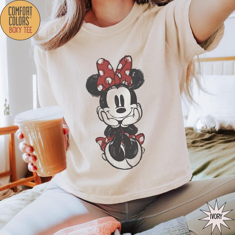 May include: Ivory-colored Comfort Colors Boxy Tee featuring a distressed Minnie Mouse graphic. The graphic shows Minnie Mouse with a red polka dot bow and matching accents. The shirt is a relaxed fit.