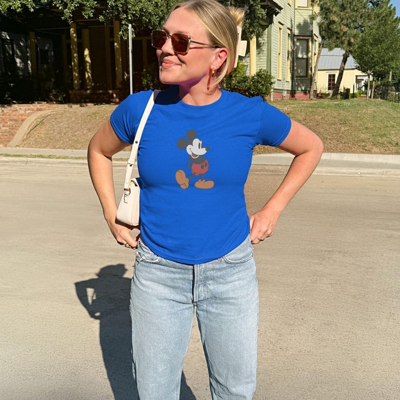 May include: A royal blue t-shirt featuring a graphic of Mickey Mouse in his classic pose. The shirt is paired with light wash denim jeans. A white purse is worn over the shoulder.