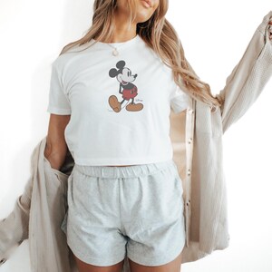 May include: White t-shirt featuring a classic Mickey Mouse graphic. The cartoon character is depicted in black, red, and tan colors. The shirt is paired with gray shorts and a beige jacket.