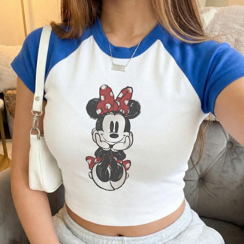 May include: White crop top with blue short sleeves featuring a Minnie Mouse graphic. The graphic shows Minnie Mouse with a red polka dot bow. The top is a casual style.
