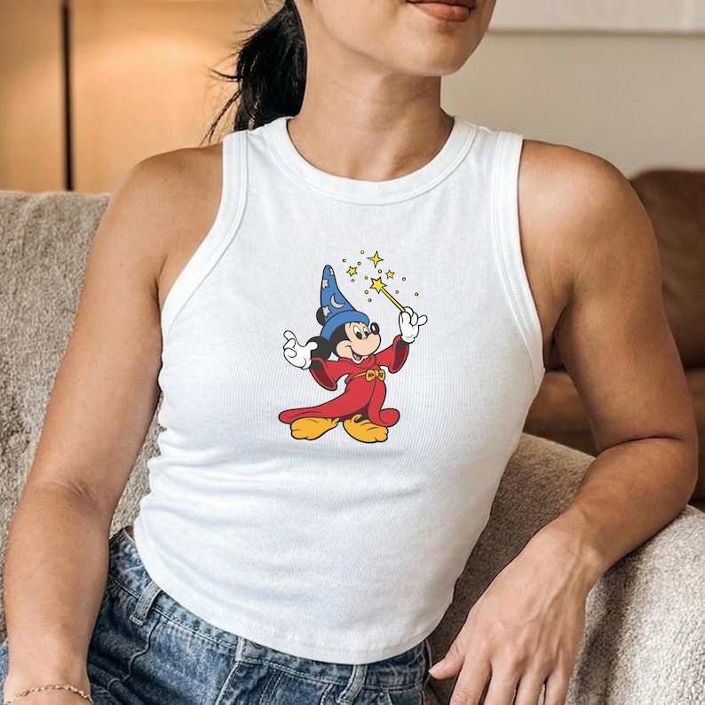 May include: White tank top featuring a colorful graphic of Mickey Mouse dressed as a wizard, holding a wand with stars. The cartoon character is wearing a blue hat, red robe, and yellow shoes. The top is sleeveless and has a ribbed texture.