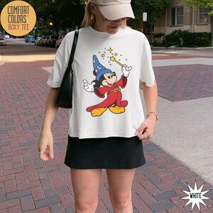 K&ouml;nnte beinhalten: Wei&szlig;es Comfort Colors Boxy T-Shirt mit einer Grafik von Mickey Mouse als Zauberer, der einen Zauberstab mit Sternen h&auml;lt. Die Zeichentrickfigur tr&auml;gt eine rote Robe und einen blauen Hut. Das Shirt hat das Wort "WHITE" unten rechts.