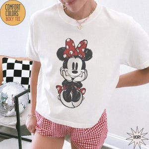 May include: Ivory-colored Comfort Colors Boxy Tee featuring a Minnie Mouse graphic. The graphic shows Minnie Mouse with a red polka dot bow and red accents. The model is wearing red and white checkered shorts.