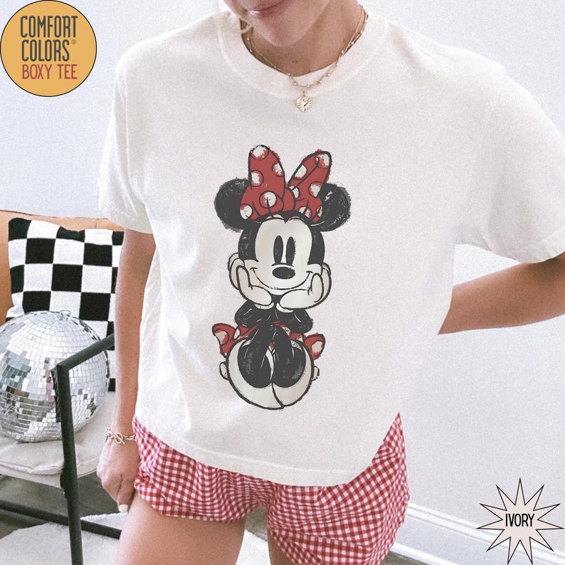 May include: Ivory-colored Comfort Colors Boxy Tee featuring a Minnie Mouse graphic. The graphic shows Minnie Mouse with a red polka dot bow and red accents. The model is wearing red and white checkered shorts.