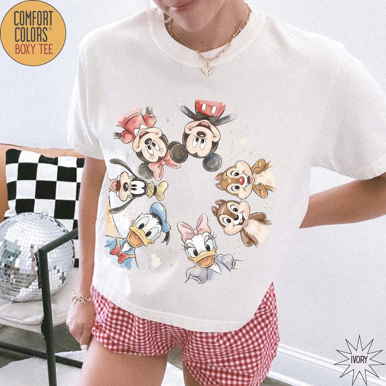 May include: Ivory-colored Comfort Colors boxy tee featuring a watercolor design of Mickey Mouse, Minnie Mouse, Donald Duck, Daisy Duck, Goofy, and Chip and Dale. The shirt has a crew neck and short sleeves. The shirt is paired with red and white checkered shorts.