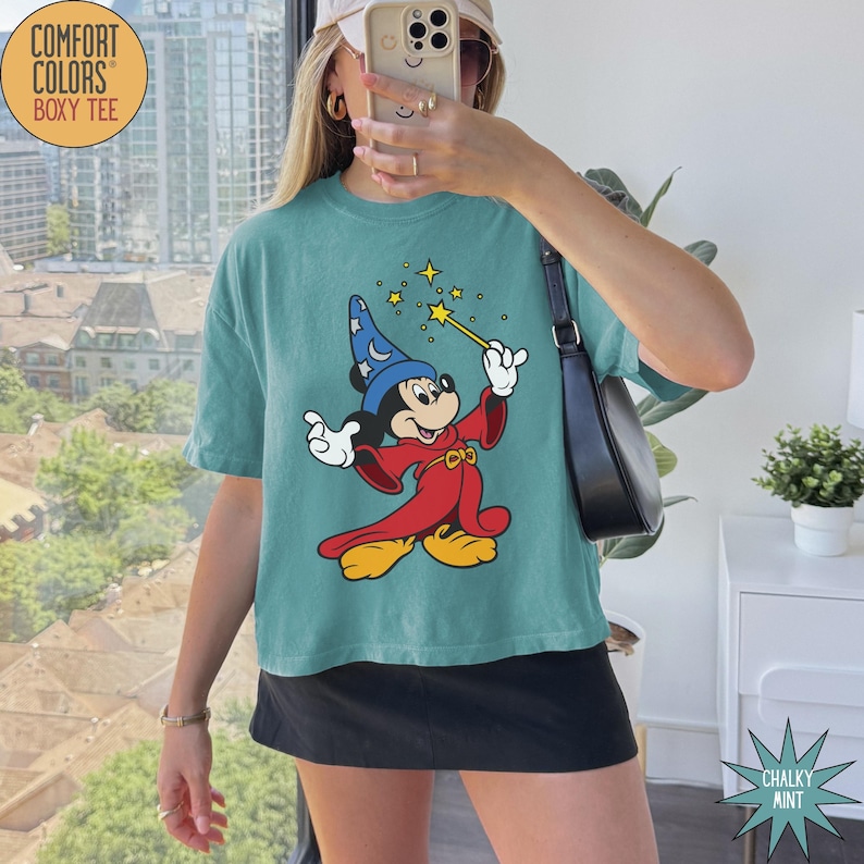 K&ouml;nnte beinhalten: T&uuml;rkisfarbenes Comfort Colors Boxy-T-Shirt mit einer Grafik von Mickey Mouse als Zauberer, der einen Zauberstab mit Sternen h&auml;lt. Die Zeichentrickfigur tr&auml;gt einen blauen Zaubererhut und eine rote Robe. Auf dem Shirt steht "Chalky Mint".