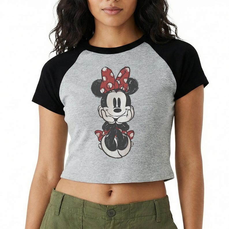 May include: A gray raglan crop top featuring a Minnie Mouse graphic. The graphic shows Minnie Mouse with a red bow with white polka dots. The sleeves are black.