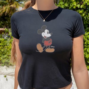 May include: Black t-shirt featuring a classic Mickey Mouse graphic. The cartoon character is depicted in his iconic pose, wearing red shorts and large brown shoes. The Disney logo is visible. A gold necklace is also visible.
