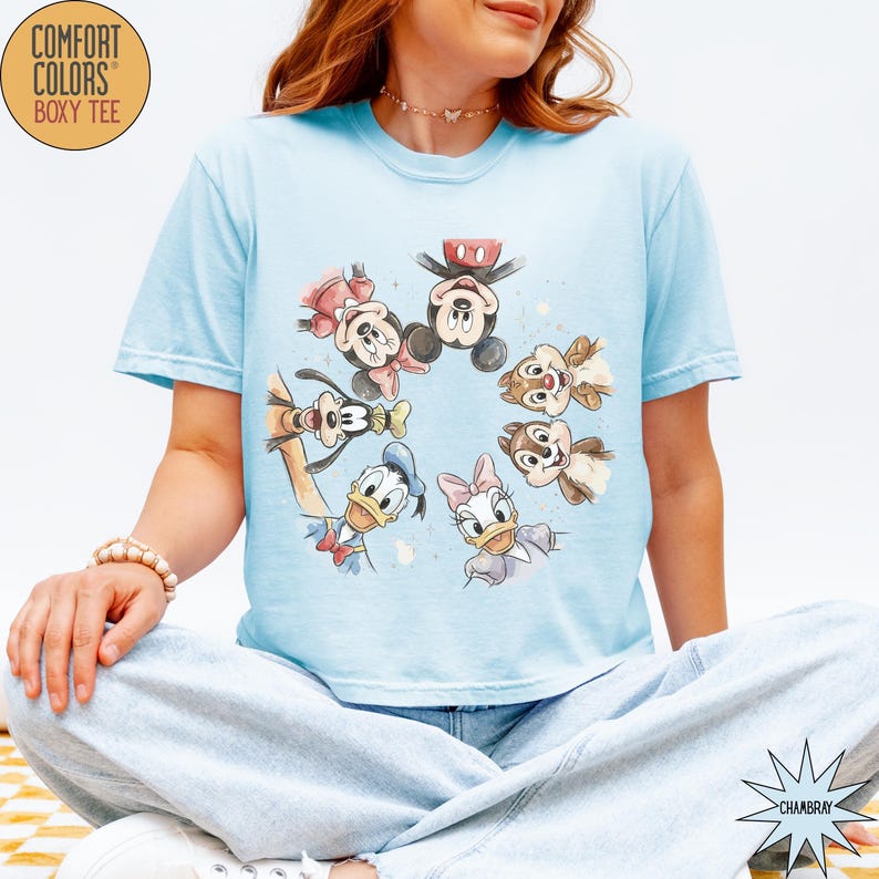 May include: Light blue Comfort Colors boxy tee featuring a watercolor design of Disney characters including Mickey Mouse, Minnie Mouse, Donald Duck, Daisy Duck, Goofy, and Chip and Dale. The shirt has short sleeves.