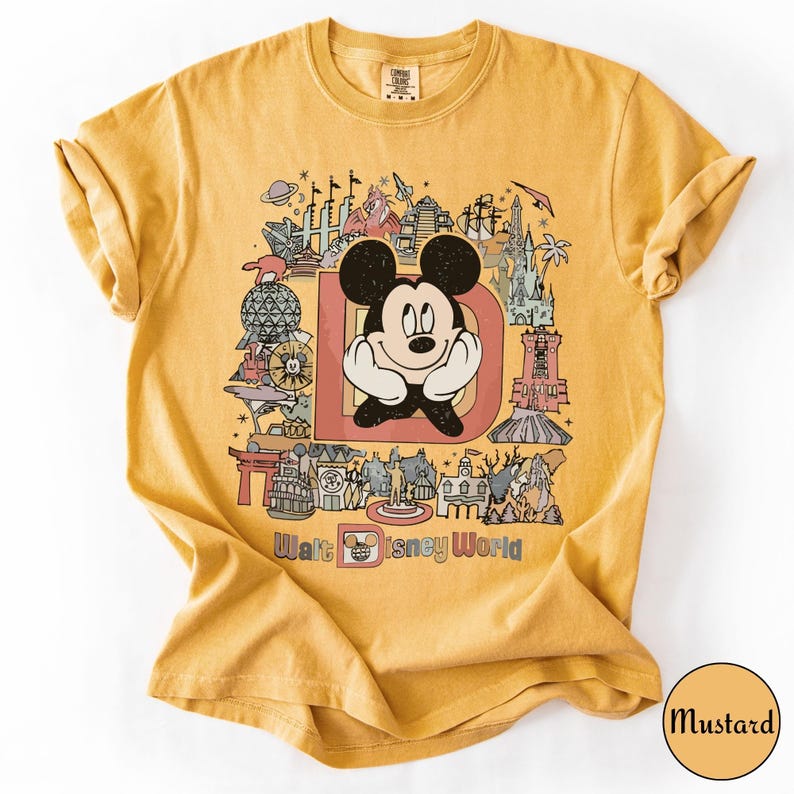 Puede incluir: Camiseta de color mostaza con un gr&aacute;fico de Mickey Mouse dentro de una letra "D" y varios monumentos de Disney World. Las palabras "Walt Disney World" est&aacute;n impresas debajo. La camiseta es de material suave.