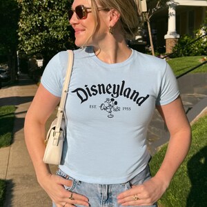 May include: Pale blue t-shirt featuring the word "Disneyland" in black script, a Mickey Mouse graphic, and the text "EST. 1955". The top has short sleeves and a fitted design.