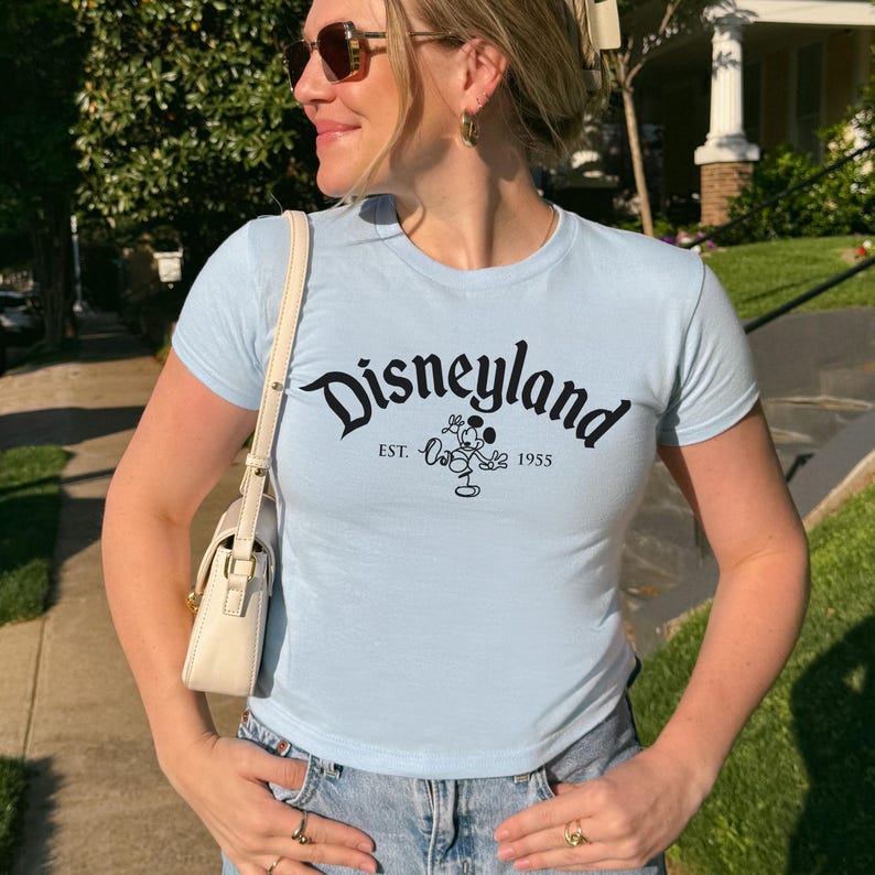 May include: Pale blue t-shirt featuring the word "Disneyland" in black script, a Mickey Mouse graphic, and the text "EST. 1955". The top has short sleeves and a fitted design.