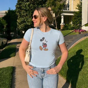 May include: Light blue t-shirt featuring a classic Mickey Mouse graphic. The shirt has short sleeves and a crew neck. The person is wearing blue jeans and sunglasses. A white purse is hanging from the shoulder.