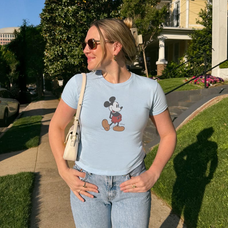 May include: Light blue t-shirt featuring a classic Mickey Mouse graphic. The shirt has short sleeves and a crew neck. The person is wearing blue jeans and sunglasses. A white purse is hanging from the shoulder.