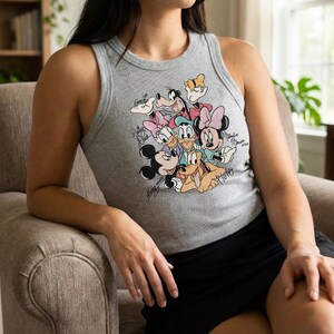 K&ouml;nnte beinhalten: Graues geripptes Tanktop mit einem farbenfrohen Aufdruck von Disney-Figuren, darunter Micky Maus, Minnie Maus, Donald Duck und Goofy. Das Tanktop hat einen Rundhalsausschnitt und ist &auml;rmellos. Der Aufdruck enth&auml;lt Unterschriften der Charaktere.