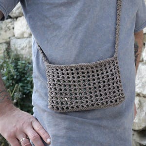 May include: A small, open-weave, brown crochet crossbody bag. The bag is rectangular with a long strap. The bag is worn over the shoulder. The background is a stone wall and greenery.