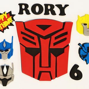 May include: A selection of colourful Transformers-themed decorations. It includes a large red and black Autobot symbol, character heads, the name "RORY" in black letters, a number "6", and a "Happy Birthday" graphic.