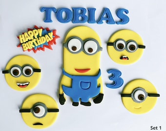 May include: A collection of yellow Minion-themed cake decorations. Includes a large Minion figure, several round Minion faces, the name "TOBIAS" in blue letters, the number "3", and a "HAPPY BIRTHDAY" accent.