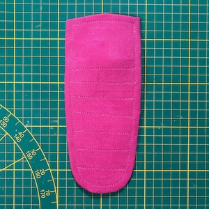 May include: A magenta-colored, rectangular fabric item with rounded edges, featuring horizontal stitched lines. The item is placed on a green cutting mat with grid lines and measurement markings.