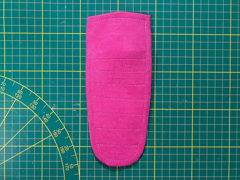 May include: A magenta-colored, rectangular fabric item with rounded edges, featuring horizontal stitched lines. The item is placed on a green cutting mat with grid lines and measurement markings.