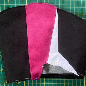 May include: A partially constructed black fabric item with a magenta stripe down the center and a white section on the right. Pins secure the fabric pieces on a green cutting mat. The item appears to be a hat or head covering.