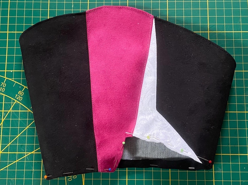 May include: A partially constructed black fabric item with a magenta stripe down the center and a white section on the right. Pins secure the fabric pieces on a green cutting mat. The item appears to be a hat or head covering.