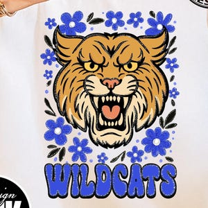 Puede incluir: Camiseta blanca con una ilustraci&oacute;n de un gato mont&eacute;s, rodeada de flores azules. La palabra "WILDCATS" est&aacute; escrita en azul con una fuente retro. El dise&ntilde;o incluye el texto "PNG | DESCARGA DIGITAL".