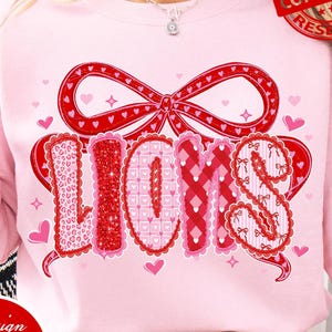 May include: A light pink sweatshirt with a large red bow and the word "LOVES" in a decorative font. The letters are in various patterns and colors, including leopard print and glitter. Small pink hearts are scattered around the design.